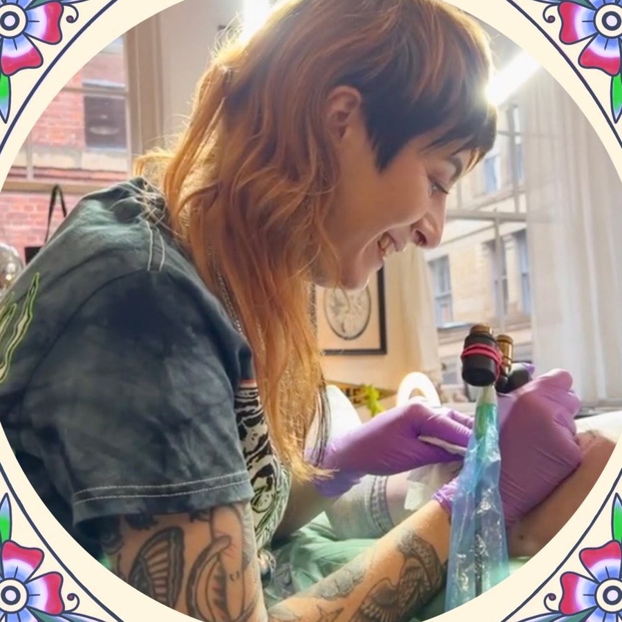 Gem Hawk tattooing a client, smiling, machine in hand